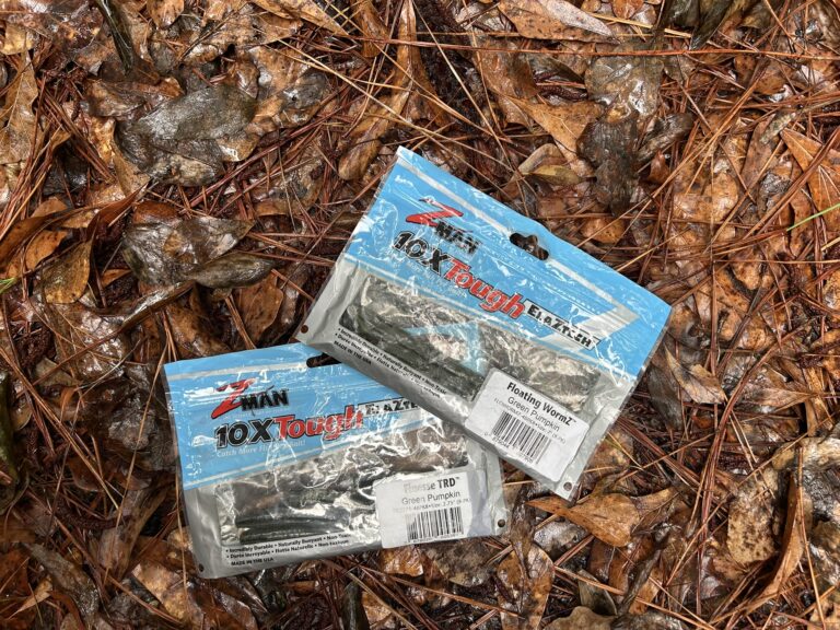 Z-Man 10X Tough ElaZtech soft plastic fishing lures in Green Pumpkin color, including Finesse TRD and Floating WormZ, photographed on wet forest ground with pine needles and autumn leaves.